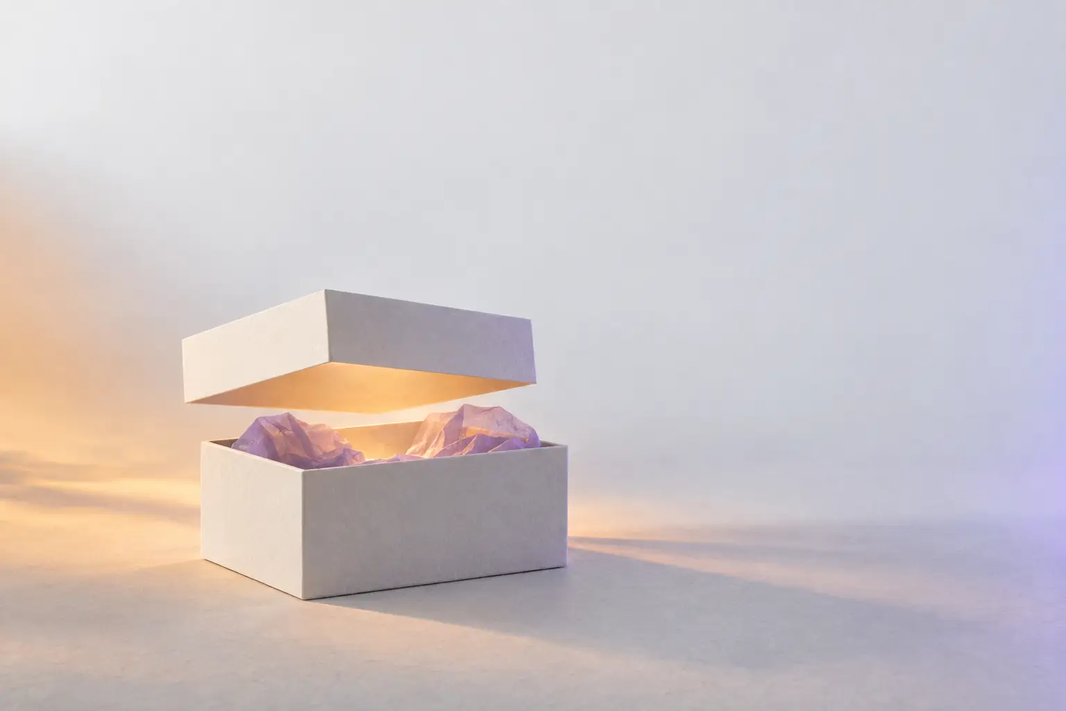 A plain off-white card box on a pale surface with its lid floating just above it, warm light spilling from inside the violet-lined interior.