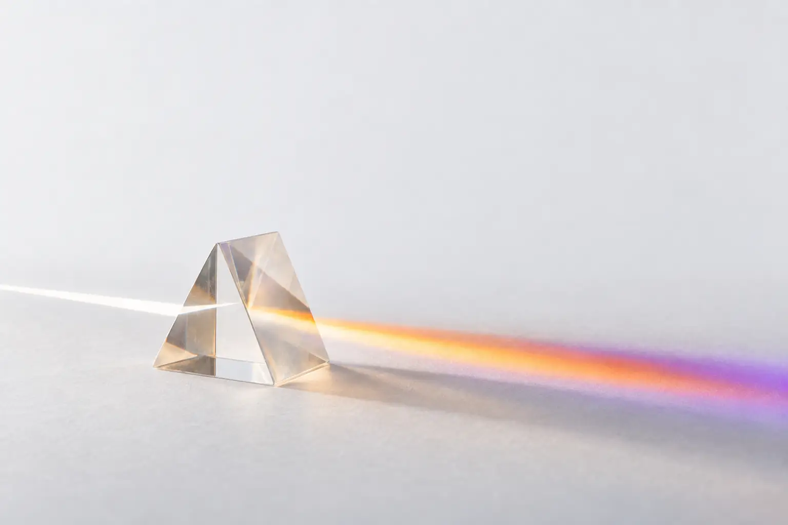 A clear glass prism on a pale surface splitting a single beam of white light into an ordered band of amber, orange and violet.