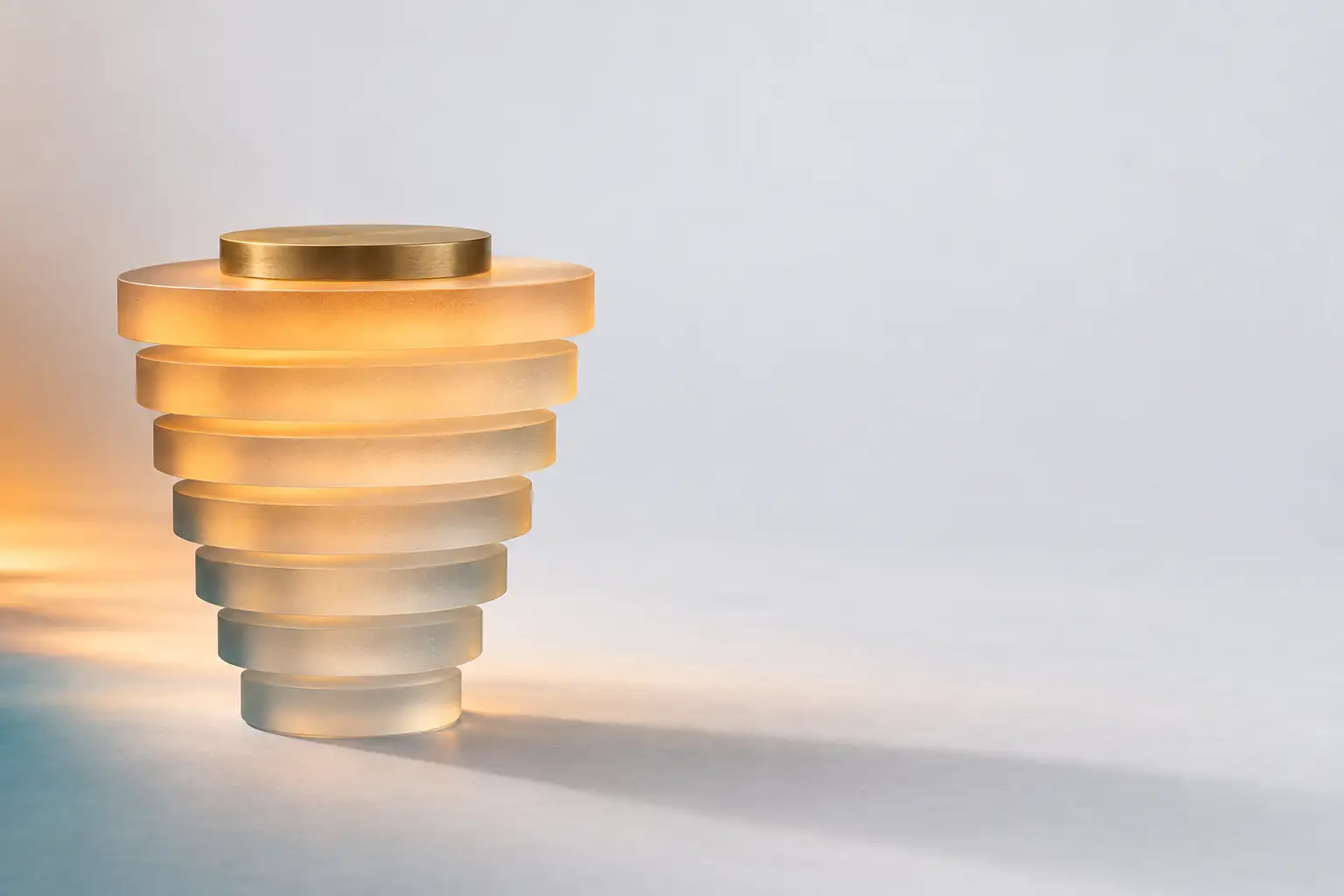 A stack of nine frosted glass discs widening as it rises, warm light travelling up through the glass, topped by a slim brass disc.