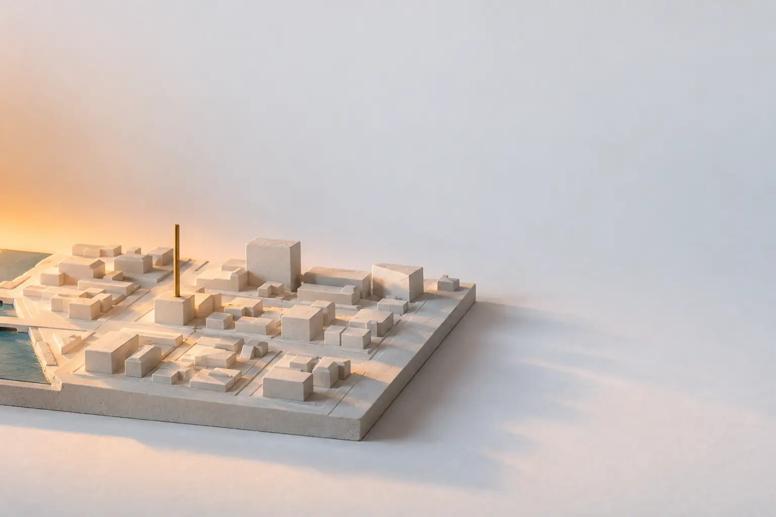 A low-relief plaster model of a town street grid with one tall brass pin standing in a single block, casting a long shadow across the model.