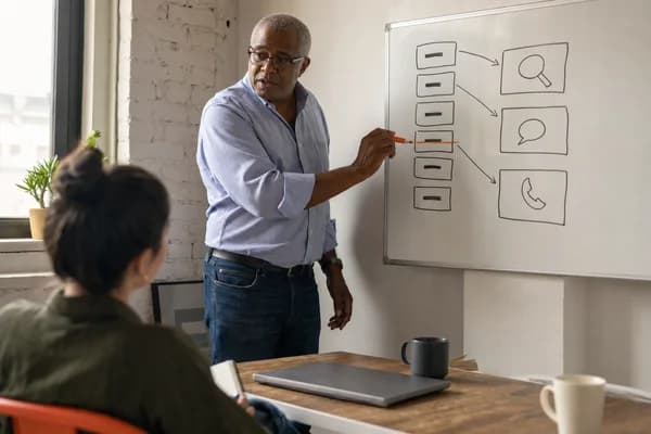 A grey-haired man in glasses and a light blue shirt strikes an orange line through one box of a hand-drawn plan of boxes and arrows on a whiteboard, while a colleague seen from behind listens with a notebook on her knee.