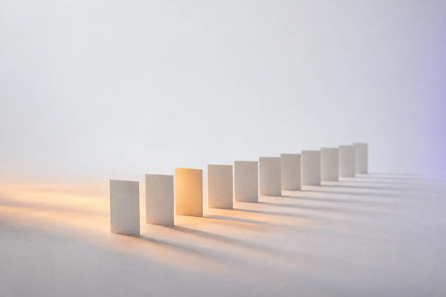 A long evenly spaced row of identical pale cards standing on edge, with one card lifted slightly proud of the line and catching warm light.